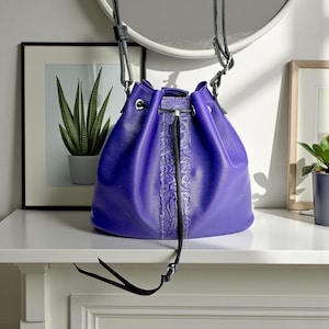 May include: A vibrant purple bucket bag with a drawstring closure and a black shoulder strap. The bag features a central embossed design and is displayed on a white surface, with a framed picture and a mirror in the background.