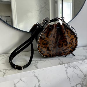 May include: A bucket bag featuring a brown and black leopard print fur-textured front panel, black leather sides, and a drawstring closure. The bag includes a long, adjustable black leather shoulder strap with silver-toned metal hardware.