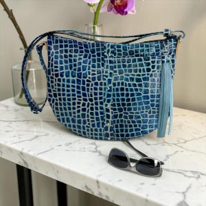 May include: A crescent-shaped shoulder bag in a deep blue crocodile-embossed pattern with a vertical center strip featuring silver-toned metal studs. The bag includes a matching blue leather tassel charm and an adjustable shoulder strap.