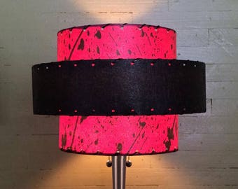 May include: A two-tiered lampshade with a black middle band and a pink top and bottom with a splatter paint design. The lampshade is made of fabric and has a stitched edge.