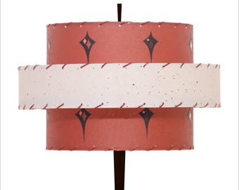 May include: A mid-century modern lamp with a tiered shade. The top and bottom sections are a reddish-orange color with black diamond designs. A white band with black speckles and red stitching accents the lamp. The lamp has a dark brown wooden base.