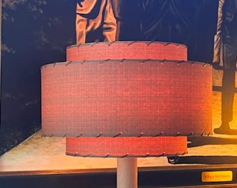 May include: A two-tiered lampshade with a brown fabric exterior and a stitched seam around the edges. The lampshade is illuminated and casts a warm glow. The lampshade is in front of a framed picture with the text "A Night With Saints".