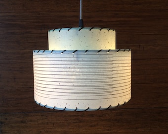 May include: A white, two-tiered pendant light with a black cord and a black trim around the edges of the shades. The top shade is a solid white, while the bottom shade has a striped pattern.