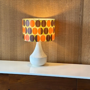 Puede incluir: Lámpara de mesa retro con base de cerámica blanca y pantalla estampada. La pantalla tiene un diseño repetido de círculos naranjas, marrones y amarillos. La lámpara está sobre una superficie blanca, con una pared de madera panelada al fondo.