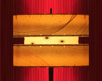 May include: A mid-century modern lamp with a rectangular shade. The shade is a warm yellow with decorative lines and starburst designs. The lamp is set against a red background.