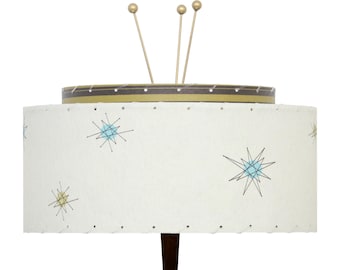 May include: A mid-century modern lamp with a white shade featuring blue and gold starburst designs. The lamp has a brown base and gold accents, including three antenna-like rods extending from the top. The shade is approximately 12 inches in diameter.