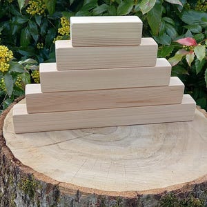 Könnte beinhalten: Ein Stapel aus fünf naturbelassenen Holzblöcken, die in Pyramidenform auf einem Baumstumpf angeordnet sind. Die Blöcke sind unbehandelt und haben eine glatte Oberfläche.