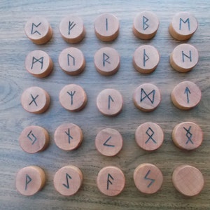 May include: Twenty-four wooden rune tokens with black ink markings. The runes are arranged in four rows of six tokens each. The runes are on a wooden surface.