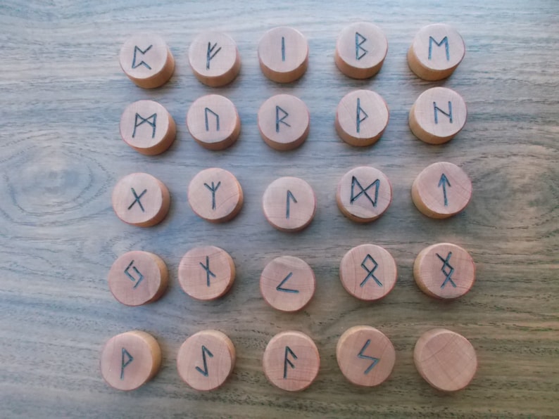 May include: Twenty-four wooden rune tokens with black ink markings. The runes are arranged in four rows of six tokens each. The runes are on a wooden surface.