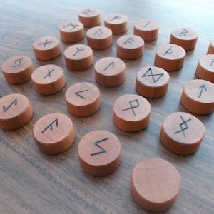 May include: A set of 24 wooden rune tokens with different symbols carved into them. The runes are arranged in a circle on a wooden surface.