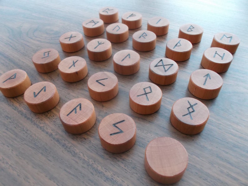 May include: A set of 24 wooden rune tokens with different symbols carved into them. The runes are arranged in a circle on a wooden surface.