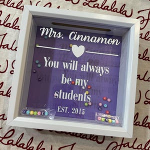 Può includere: Una scatola ombra con cornice bianca e sfondo viola. Il testo recita "Mrs. Cinnamon, You will always be my students, EST. 2015." Sono visibili anche un cuore bianco e perline colorate. Due piccoli sacchetti di perline compongono dei nomi.