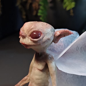 Peut inclure: Une sculpture détaillée d'une créature humanoïde à la peau pâle, avec de grands yeux rouges et des oreilles pointues. La figurine a une petite bouche et semble avoir des ailes translucides. La sculpture est finement détaillée, montrant la texture de la peau et la forme de la créature.