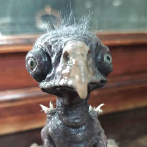 May include: A close-up of a dark gray, bird-like sculpture with a pale beak and large, round eyes. The sculpture has a tuft of light gray hair on its head and small, white appendages. The background is blurred.