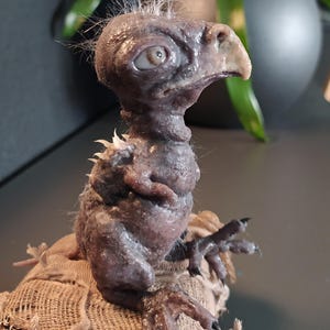May include: A detailed sculpture of a small, avian-like creature with a textured, purplish-gray skin. It has a beak, large eye, and tufts of hair. The creature is perched on a burlap sack, showcasing intricate details and a unique aesthetic.