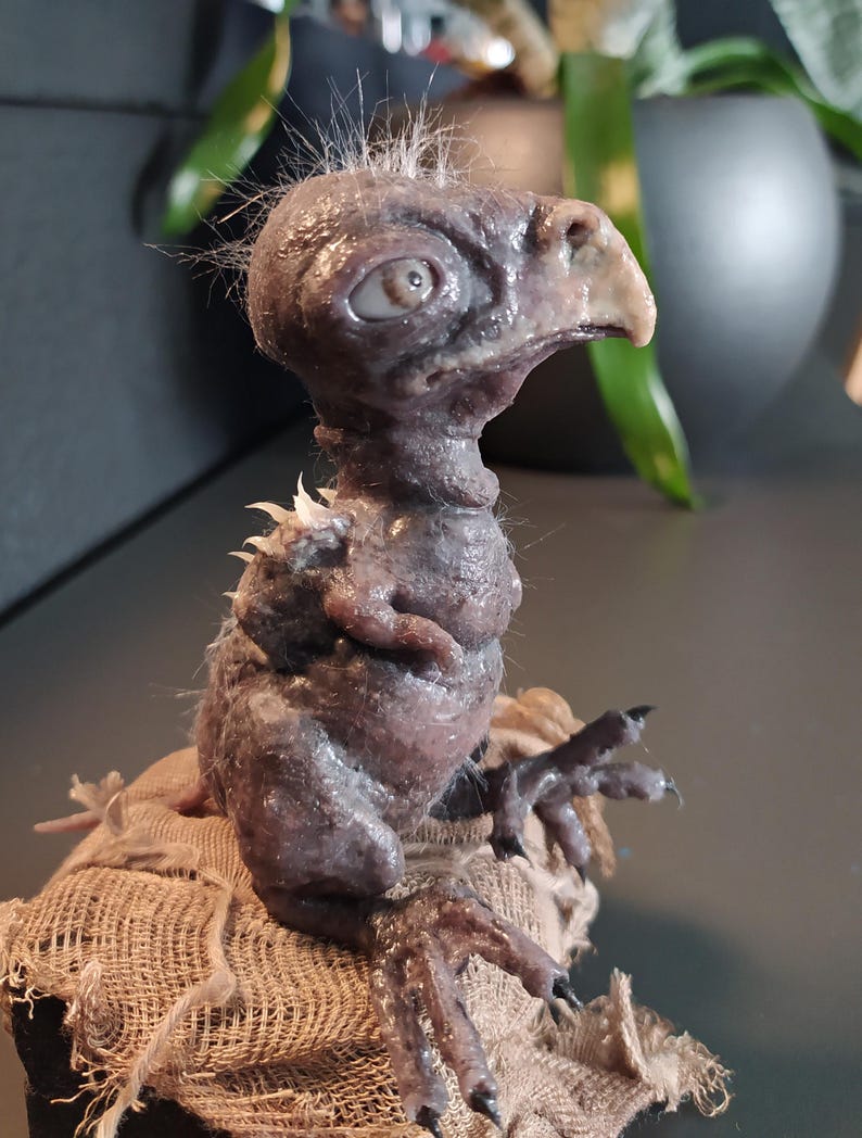 May include: A detailed sculpture of a small, avian-like creature with a textured, purplish-gray skin. It has a beak, large eye, and tufts of hair. The creature is perched on a burlap sack, showcasing intricate details and a unique aesthetic.