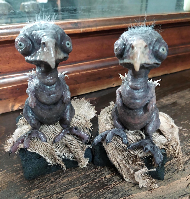 May include: Two dark gray avian sculptures with large eyes and beaks, perched on burlap-covered bases. The sculptures have a textured appearance and are set against a wooden background. The overall theme is fantasy or gothic.