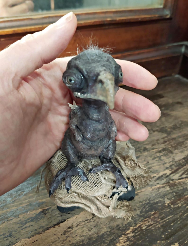 May include: A dark gray, fantastical bird-like sculpture with large eyes and a beak. The creature is perched on a piece of burlap. The sculpture has a textured appearance and is held in a hand.