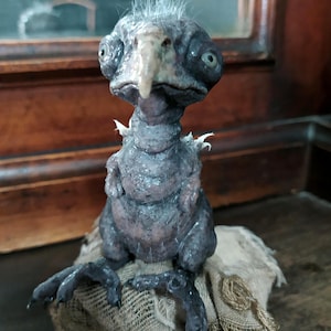 May include: A detailed sculpture of a fantastical creature with a bird-like beak and large eyes. The figure is primarily dark gray with textured skin and a tuft of white hair. It is perched on a piece of rough, tan fabric.
