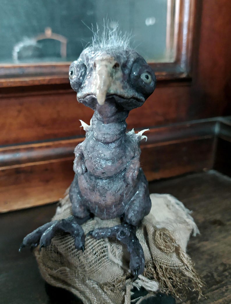 May include: A detailed sculpture of a fantastical creature with a bird-like beak and large eyes. The figure is primarily dark gray with textured skin and a tuft of white hair. It is perched on a piece of rough, tan fabric.
