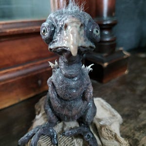 May include: A dark gray, grotesque bird-like sculpture with a long beak and large, bulging eyes. The creature has a textured, almost reptilian skin and is perched on a rough, tan fabric. The sculpture has a unique, unsettling aesthetic.