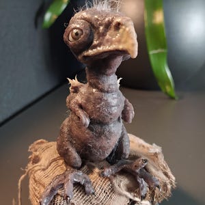 May include: A detailed sculpture of a fantastical creature with a bird-like beak and large, expressive eyes. The figure is a dark, muted brown color and sits atop a rough, textured fabric base. The sculpture has a unique, handcrafted appearance.