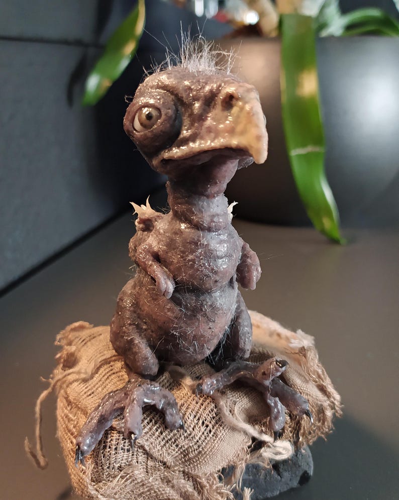 May include: A detailed sculpture of a fantastical creature with a bird-like beak and large, expressive eyes. The figure is a dark, muted brown color and sits atop a rough, textured fabric base. The sculpture has a unique, handcrafted appearance.