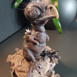 May include: A detailed sculpture of a creature with a bird-like beak, large eye, and textured, dark brown skin. The figure has small arms and is perched on a burlap sack. White wisps of hair sprout from the top of its head.