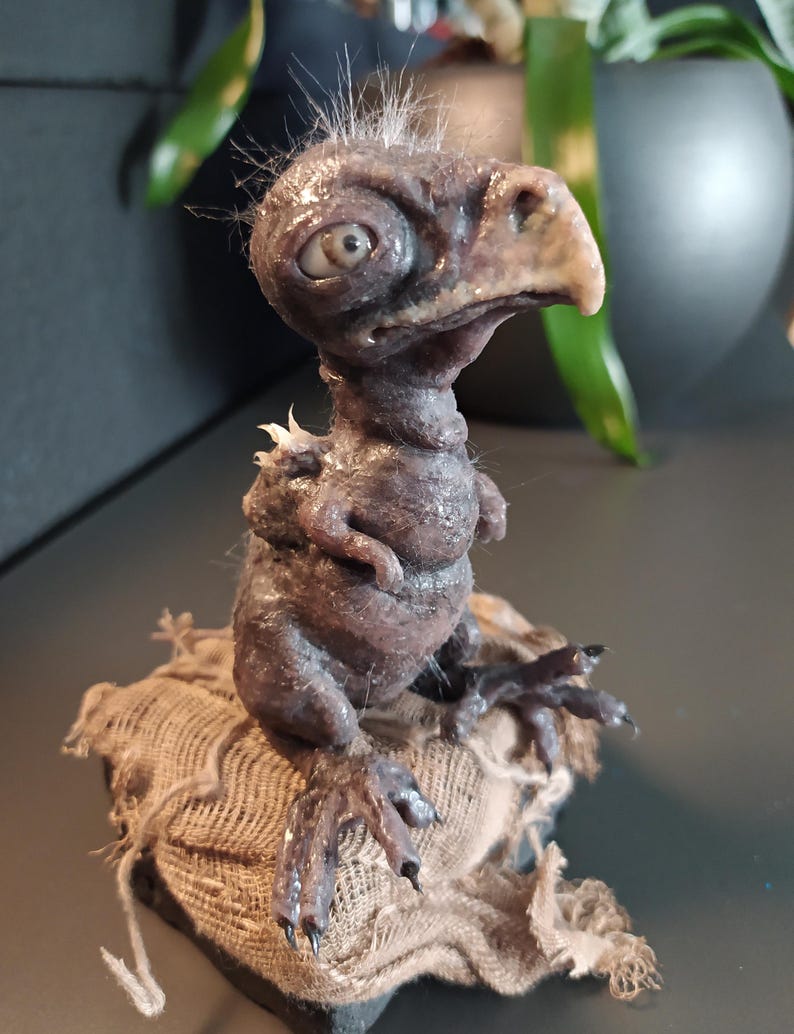 May include: A detailed sculpture of a creature with a bird-like beak, large eye, and textured, dark brown skin. The figure has small arms and is perched on a burlap sack. White wisps of hair sprout from the top of its head.