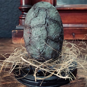 Könnte beinhalten: Eine strukturierte, eiförmige Skulptur in gedämpftem Grün mit dunkelgrauen und schwarzen Akzenten, mit einer rissigen Oberfläche. Sie ruht auf trockenem Gras, auf einem schwarzen, kreisförmigen Sockel. Ein einzigartiges Dekorationsobjekt.