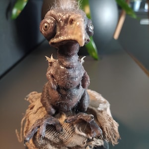 May include: A detailed sculpture of a fantastical creature with a bird-like beak and large eyes. The figure is a dark brown color with a textured surface and sits atop a piece of rough, tan fabric. The sculpture has small, spiky protrusions.