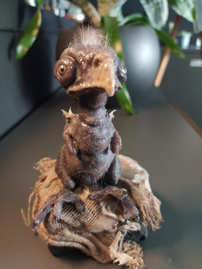 May include: A detailed sculpture of a fantastical creature with a bird-like beak and large eyes. The figure is a dark brown color with a textured surface and sits atop a piece of rough, tan fabric. The sculpture has small, spiky protrusions.