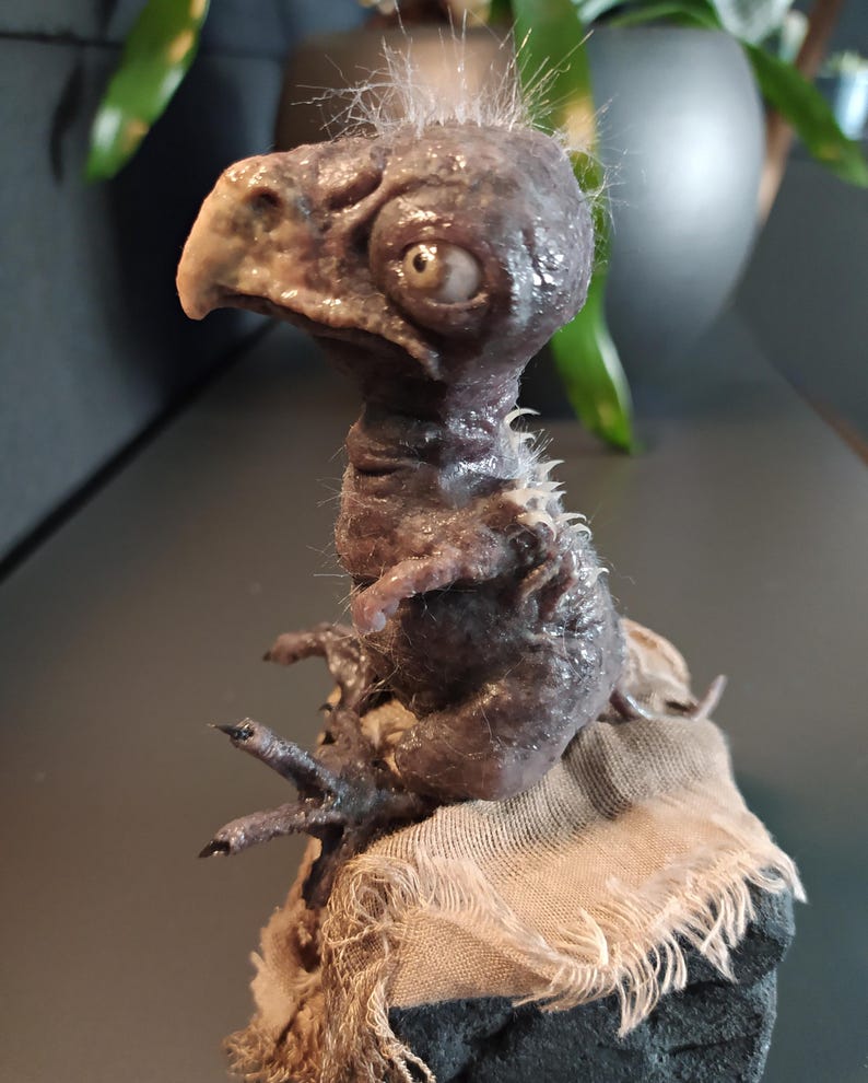May include: A detailed sculpture of a creature with a bird-like beak, large eye, and textured skin. The figure is primarily dark brown with hints of lighter tones, and sits atop a rough, tan fabric. Fine, white hairs sprout from the head.