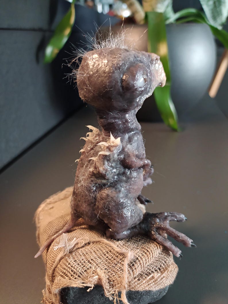 May include: A dark, textured sculpture of a creature with a large head, small body, and clawed hands. The sculpture is dark purple and gray, with a rough, organic appearance. It sits on a small, burlap-covered base.