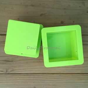 Könnte beinhalten: Hellgrüne quadratische Silikonform mit abgerundeten Ecken, neben einem Stapel ähnlicher Formen. Der Text "Daily Proposal" ist auf der Seite der Form sichtbar. Die Formen befinden sich auf einer Holzoberfläche.