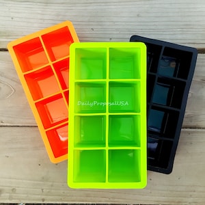 May include: Three silicone ice cube trays in orange, green, and black. Each tray has eight square molds. The green tray has the text "DailyProposalUSA" printed on it. The trays are arranged on a wooden surface.