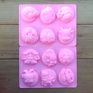 Könnte beinhalten: Zwei rosa Silikonformen mit jeweils sechs Tierkreiszeichen-Designs. Die Zeichen umfassen Widder, Stier, Zwillinge, Krebs, Löwe, Jungfrau, Waage, Skorpion, Schütze, Steinbock, Wassermann und Fische. Für Seifen oder andere Bastelarbeiten.