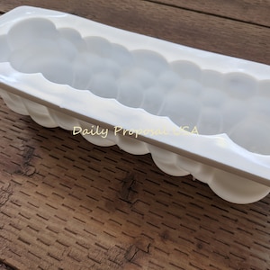 May include: A white, rectangular mould with a cloud-like design. The mould has a series of rounded, bubble-like shapes along its sides and interior. The text "Daily Proposal USA" is visible on the surface. The mould is on a wooden surface.
