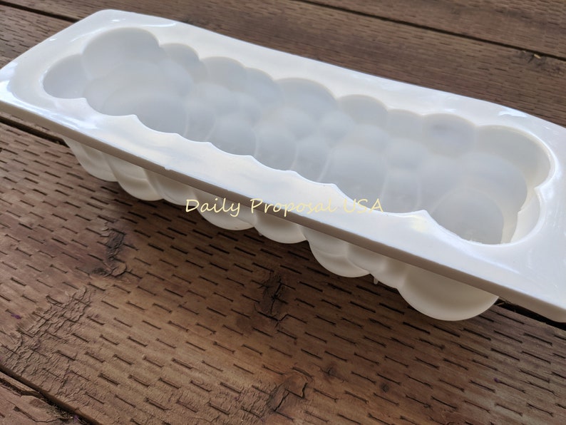 May include: A white, rectangular mould with a cloud-like design. The mould has a series of rounded, bubble-like shapes along its sides and interior. The text "Daily Proposal USA" is visible on the surface. The mould is on a wooden surface.