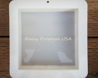 May include: A white square mold with rounded corners and a small hole at the top. The text "Daily Proposal USA" is printed in a light yellow color in the center of the mold. The mold is sitting on a wooden surface.