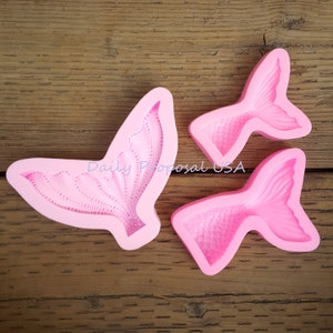 May include: Pink silicone molds in the shape of mermaid tails. One mold is a large, open tail design, and the other two are smaller, closed tail designs. The molds are on a wooden surface.