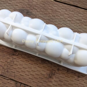 May include: A white ice mould with a series of rounded compartments, designed to create spherical ice shapes. The words "Daily Proposal USA" are printed on the side. The mould sits on a wooden surface.