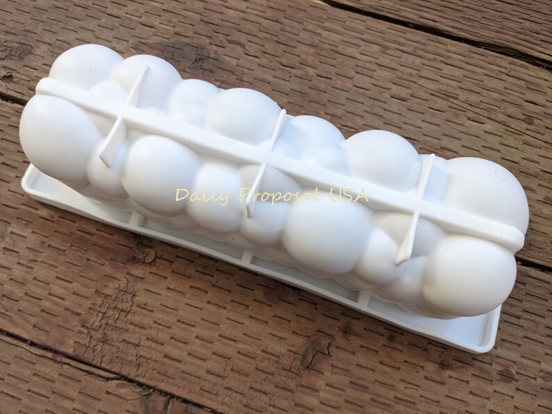 May include: A white ice mould with a series of rounded compartments, designed to create spherical ice shapes. The words "Daily Proposal USA" are printed on the side. The mould sits on a wooden surface.
