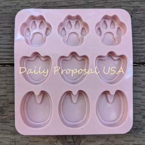 May include: A light pink silicone mold with nine cavities. The cavities are shaped as three paw prints, three duck feet, and three horse hooves. The text "Daily Proposal USA" is printed on the mold.