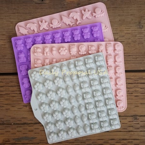 Può includere: Tre stampi in silicone rosa, viola e grigio, ciascuno pieno di varie forme. Gli stampi presentano disegni di animali e creature marine, tra cui orsi, delfini e stelle marine. Il testo "Daily Proposal USA" è visibile.
