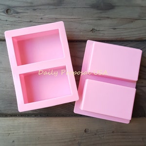 May include: Two pink silicone soap molds. One mold has two rectangular cavities, and the other has two rectangular cavities. The text "Daily Proposal USA" is visible on the mold with two cavities.