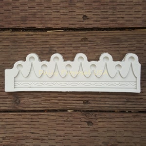 May include: A white silicone crown mold with a detailed design. The mold features a series of rounded peaks with teardrop-shaped accents and a decorative lower border. The text "Daily Proposal USA" is visible on the mold.