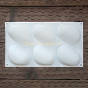 Puede incluir: Un molde de silicona blanco con seis cavidades de forma ovalada. El molde está sobre un fondo de madera. El texto "Daily Proposal US" está impreso en amarillo en el centro del molde.