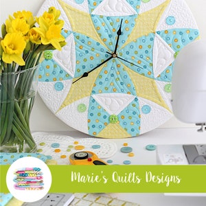 May include: A quilted clock with a blue, yellow, and white design. The clock is surrounded by buttons and a sewing machine. The text "Quilt Clock" is at the top of the image and "Marie's Quilts Designs" is at the bottom.