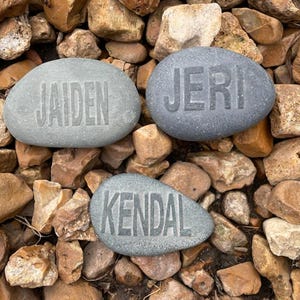 May include: Three gray painted rocks with the names Jaiden, Jeri, and Kendal written on them. The rocks are sitting on a bed of brown and gray rocks.